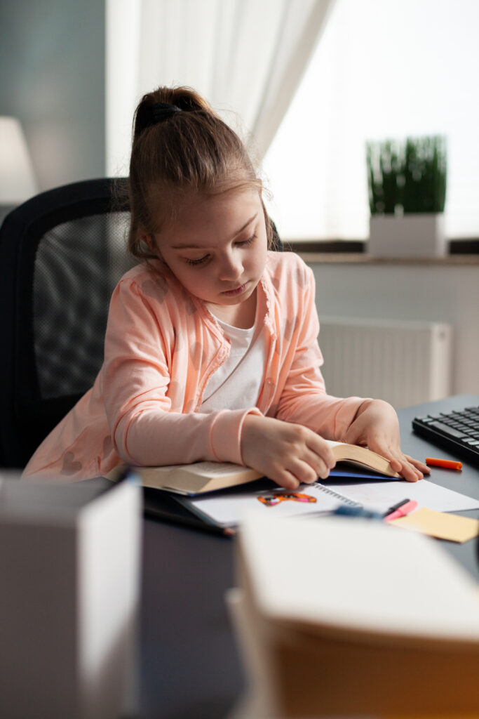 Schoolchild sitting at desk table in living room holding homework book reading educational literature story for online course during coronavirus quarantine. Reader child studying for school exam